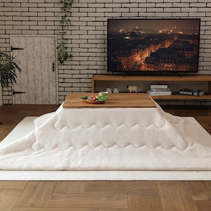May include: A white electric heated blanket with a quilted pattern, placed on a wooden table with a plate of food. The table is in front of a brick wall with a white door and a television playing a cityscape at night.