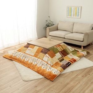 Puede incluir: Una mesa baja de madera clara está colocada sobre una gran colcha de retazos colorida. La colcha presenta una variedad de patrones en tonos naranja, marrón, verde y crema. Debajo de la colcha hay una alfombra beige.