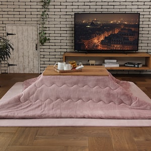 May include: A pink electric heated blanket with a quilted design sits on a wooden table with two mugs and a tray. The blanket is on a light pink rug in front of a brick wall with a TV and a shelf with books and a small sheep figurine.
