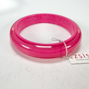 May include: A bright pink bangle bracelet. The glossy surface reflects light, enhancing the colour. A white tag with the brand name "zsiska" is attached. This accessory is a striking statement piece.