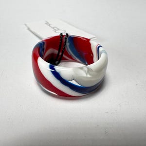 Może przedstawiać: Pierścień z wzorem czerwono-biało-niebieskiego wiru. Pierścień wykonany jest z gładkiego, błyszczącego materiału.