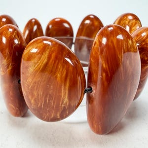 May include: A chunky bracelet featuring large, polished, oval-shaped beads in shades of brown and amber. The beads are strung together with a black cord, creating a bold and eye-catching accessory. The bracelet is a statement piece.