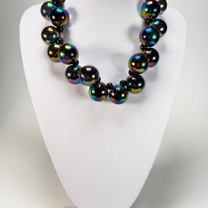 May include: A statement necklace featuring large, iridescent, round beads in shades of purple, green, and gold. The beads are strung together with smaller black beads. The necklace is displayed on a white mannequin bust.