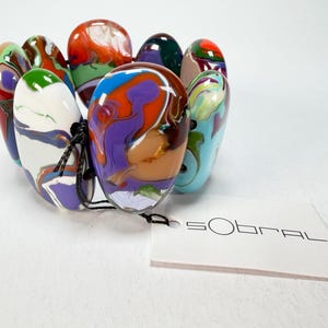 May include: A colorful bracelet featuring oval-shaped beads in various colors, including red, green, purple, and blue. The beads have a marbled effect and are strung together with a black cord. A white tag with the brand name "SOBRAL" is attached.