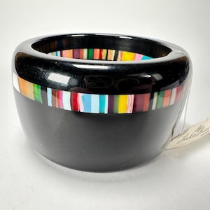 May include: Black bangle bracelet with a colorful stripe design. The bracelet is made of resin and has a glossy finish.