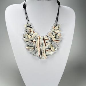 May include: A statement necklace with a unique design. The necklace features a series of abstract, layered shapes in shades of white, silver, and brown, strung on a black cord. The necklace is displayed on a white mannequin.