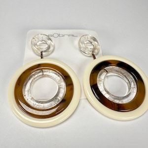 May include: A pair of large, round, cream-colored earrings with a brown and clear layered design. The earrings have a smaller clear circle inside the larger circle.