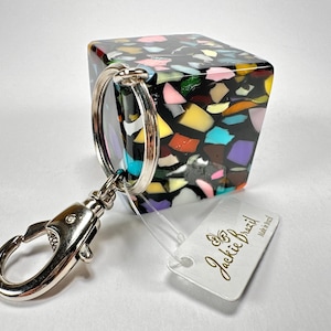 May include: A square keychain charm with a colourful terrazzo design. The charm is attached to a silver keyring and a lobster clasp. A white tag with the text "Jackie Brazil" is attached.