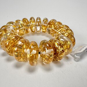 May include: A clear, spiral hair tie with gold leaf flakes encased within each bead. The beads are a translucent amber color. A white tag is attached to the hair tie.