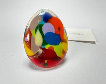 May include: A colorful, abstract glass ring with a swirl of red, orange, yellow, green, blue, and purple. The ring is oval shaped and has a clear glass base.