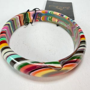 May include: A colorful, striped bangle bracelet with a variety of colors including pink, green, yellow, and brown. The bracelet has a smooth, glossy finish and a circular shape. A tag is attached to the bracelet.