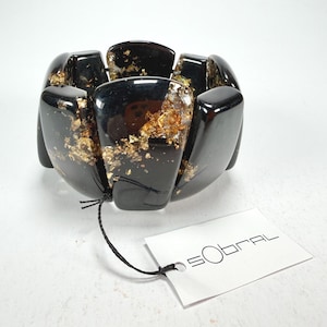 May include: A black bracelet with gold leaf accents. The bracelet is made of several curved, glossy black segments, each with gold leaf embedded within. A white tag with the brand name "sobral" is attached.