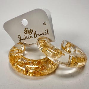 May include: Clear, C-shaped earrings with gold leaf flakes suspended within the resin. The earrings are displayed with a white tag that reads "Jackie Brazil" in a script font. The earrings are a unique jewelry item.