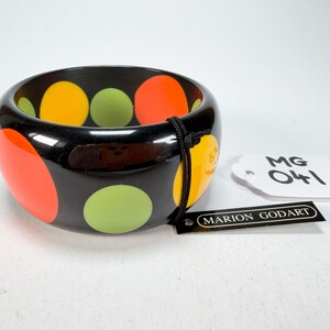 May include: A black, wide bangle bracelet with large, colourful polka dots in orange, yellow, and green. The bracelet has a glossy finish and a tag that reads "MARION GODART".