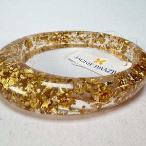 Könnte beinhalten: Ein klarer Armreif aus Harz mit Goldblatt-Einschlüssen. Das Armband hat eine runde Form und eine glatte Oberfläche. Ein weißes Etikett mit dem Text "JACKIE BRAZIL" ist angebracht.