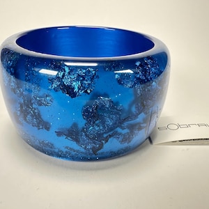 May include: A blue resin bangle bracelet with a metallic blue glitter design. The bracelet is made by the brand "Sobral".