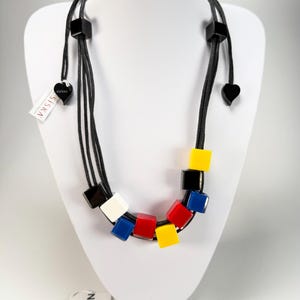 May include: A multi-strand black cord necklace featuring geometric cube beads in primary colours of red, yellow, blue, white, and black. The necklace includes small heart-shaped accents and a white tag with the text ZSISKA printed in black.