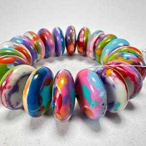 Könnte beinhalten: Ein farbenfrohes Armband mit großen, ovalen Perlen. Die Perlen zeigen eine lebendige Mischung aus Farben wie Blau, Rosa, Grün, Lila und Orange mit Marmoreffekt. Das Armband ist auf einem transparenten elastischen Band aufgefädelt.