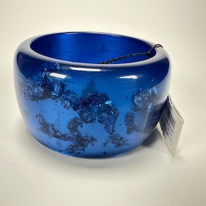 May include: A vibrant blue resin bangle bracelet. The wide, rounded bracelet features embedded dark blue metallic flakes. The bracelet has a glossy finish and a tag attached. A stylish accessory for everyday wear.