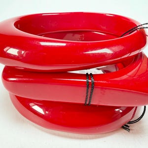 Puede incluir: Tres brazaletes rojos brillantes apilados. Los brazaletes tienen un diseño liso y redondeado. Una cuerda negra está enrollada alrededor de los brazaletes. Una etiqueta blanca está adjunta. Accesorio audaz.