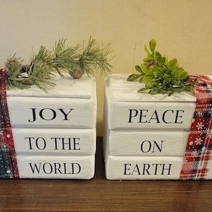 Puede incluir: Dos cajas de madera blanca apiladas una encima de la otra. La caja superior dice "JOY TO THE WORLD" y la caja inferior dice "PEACE ON EARTH". Ambas cajas están decoradas con una cinta de cuadros rojos y verdes.