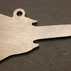 May include: A silver-colored metal pendant in the shape of a stylized map with a loop at the top for hanging. The pendant has a long, pointed extension and is set against a dark background.