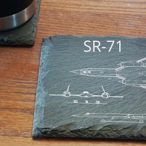 May include: A square, dark gray slate coaster with the text "SR-71" and white line drawings of the SR-71 Blackbird aircraft. The coaster has a rough, natural edge. A black tumbler sits on a similar coaster.