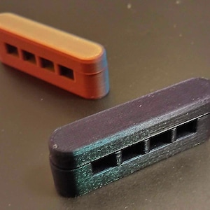 May include: Two rectangular, 3D-printed objects. One is orange, the other is black. Each has four rectangular cutouts. The objects are likely components or parts for a larger item.