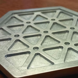 May include: A hexagonal metal coaster with a geometric design. The coaster is silver-colored and features a pattern of triangles and small circular holes. The surface has a brushed texture. The coaster is likely used for placing drinks or other items.
