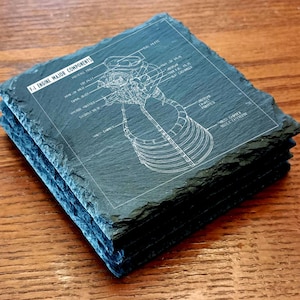 May include: A stack of four square slate coasters. Each coaster features a detailed white diagram of an F-1 engine with labels. The text "F-1 ENGINE MAJOR COMPONENTS" is visible. The coasters have a dark gray color and a rough, textured surface.