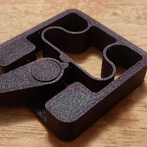 Puede incluir: Un clip de plástico gris oscuro impreso en 3D, con un marco cuadrado, detalles internos curvos y una palanca articulada. El clip está diseñado para asegurar o sujetar objetos, con una superficie texturizada.