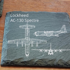 May include: A dark gray slate coaster with white line drawings of the Lockheed AC-130 Spectre aircraft. The text "Lockheed AC-130 Spectre" is printed in white. The coaster is square and sits on a wooden surface.