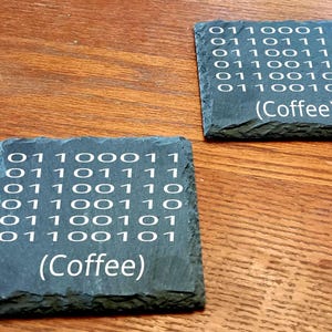 May include: Two square slate coasters with white binary code and the word "(Coffee)". The coasters are dark gray and sit on a wooden surface. A unique and tech-inspired home decor item.