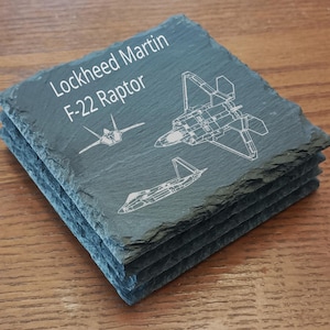 May include: A stack of four square slate coasters. Each coaster features the text "Lockheed Martin F-22 Raptor" and white line drawings of the F-22 fighter jet. The coasters have a dark gray color and a rough, textured surface.