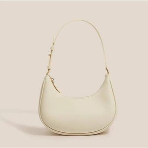 May include: A white leather handbag with a curved shape and a gold-tone zipper. The bag has a single strap.