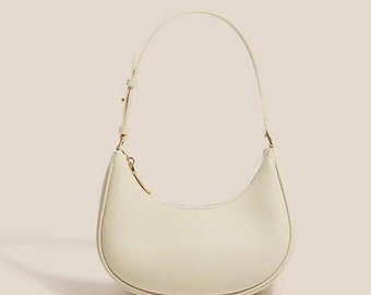 Peut inclure: Un sac à main en cuir blanc avec une forme incurvée et une fermeture éclair dorée. Le sac a une seule bandoulière.