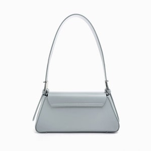 May include: A light gray shoulder bag with a rectangular body and a flap closure. The bag has a long, thin strap and silver-colored hardware. The bag is made of a smooth material and is set against a white background.