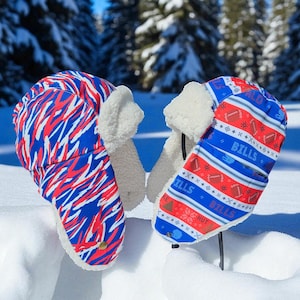 May include: Two winter hats with ear flaps, resting in the snow. One hat features a blue and red abstract pattern, while the other displays a blue background with red footballs and the word "BILLS". Both have white faux fur lining.