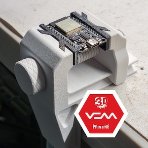 Könnte beinhalten: Ein weißer 3D-gedruckter Schraubstock, der eine Leiterplatte mit einem USB-C-Anschluss hält. Der Schraubstock hat eine strukturierte Oberfläche und eine Schraube zur Einstellung des Griffs. Die Leiterplatte hat verschiedene elektronische Komponenten. Das Bild enthält ein rot-weißes Logo mit der Aufschrift "3D VCM PRINTING".