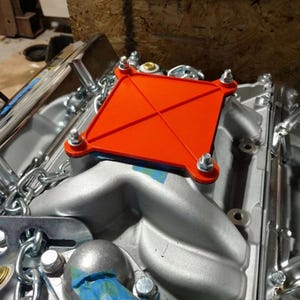 May include: An orange square component with a cross design is bolted onto a silver engine. The engine has various chrome and metal parts, including chains and bolts. The background is a wooden surface.