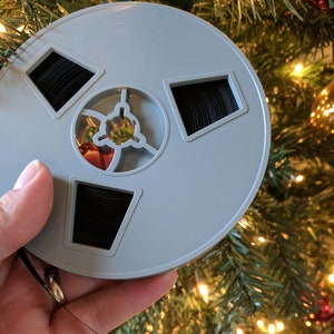 Peut inclure: Une bobine de film grise vintage avec du film noir à l'intérieur, tenue devant un sapin de Noël. La bobine a un design circulaire avec trois ouvertures rectangulaires et un moyeu central. L'arrière-plan est flou avec des lumières et des ornements de Noël.