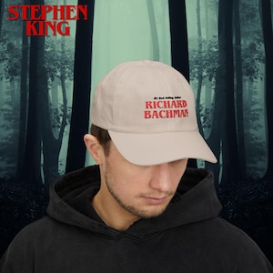 May include: A beige baseball cap with the text "#1 Best Selling Author RICHARD BACHMAN" embroidered in red. The cap has a curved brim and is made of a soft fabric. The Stephen King logo is in the top left corner.