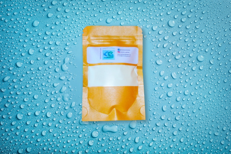 Op de afbeelding: Een kleine, bruine zak met een doorzichtig venster, gevuld met een witte substantie. De zak heeft een label met tekst en een logo. De achtergrond is een lichtblauwe ondergrond bedekt met waterdruppels.