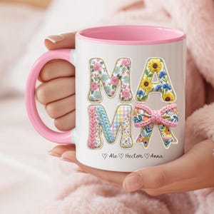 May include: White ceramic mug with a pink interior and handle. The word "MAMA" is displayed in floral and patterned letters. The mug is held by a person. The names "Ale", "Hector", and "Anna" are below the word "MAMA".