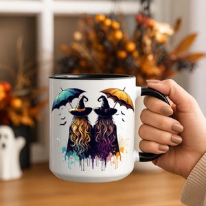 May include: A white ceramic mug with a black interior and handle. The mug features a colorful illustration of two witches with long hair, wearing hats and capes, holding umbrellas. The design includes bats and watercolor splashes, perfect for Halloween.