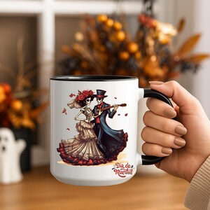 May include: White ceramic mug with a black interior and handle. The mug features a Dia de Muertos design with a skeleton couple, a woman in a dress and a man playing a guitar. The text "Dia de Muertos" is also on the mug.
