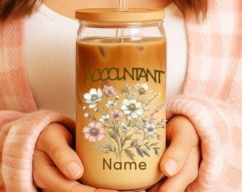 Op de afbeelding: Een heldere glazen beker met een houten deksel en rietje, gevuld met een koffiedrank. De beker is versierd met het woord "ACCOUNTANT" en een bloemenontwerp in wit, roze en groen. Het woord "Name" staat er ook op.