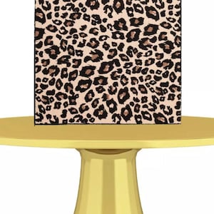 Pode incluir: Uma caixa quadrada com estampa de leopardo, com manchas marrons e pretas em um fundo bege, está em cima de um suporte para bolo amarelo. O suporte para bolo é de cor amarela sólida. A caixa tem uma borda preta.