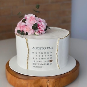 May include: A white cake with a gold trim and a floral arrangement of pink flowers. The cake has the text "AGOSTO 1994" and a calendar. The cake is on a white plate and a wooden base.