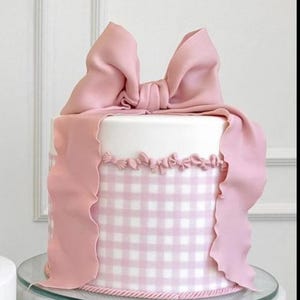Peut inclure: Un gâteau rond avec un dessus blanc et des côtés à motifs vichy roses. Le gâteau est décoré d'un grand nœud rose et d'un ruban rose qui retombe sur le côté. Le gâteau repose sur un présentoir à gâteau en verre et en marbre.